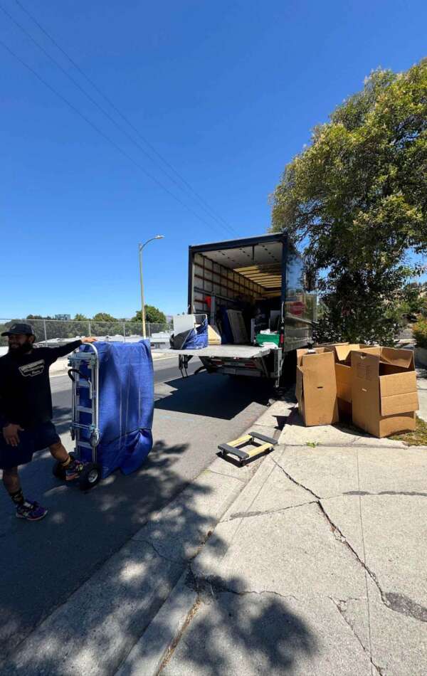 Mover with a dolly beside an open moving truck and stacked boxes on the street.